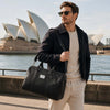 Man carrying a TL Voyager Leather Duffle Bag Small by the water with iconic buildings in the background.