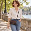 A woman wearing a cream linen shirt and blue jeans walks along a stone bridge by the water, carrying the Chocolate Crossbody Phone Wallet for Women