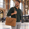 A man in a dark green polo shirt and light gray trousers carries The Natural Executive Leather Briefcase with gold-tone hardware in a large, historic train station with arched windows and a train in the background.