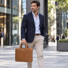 A man in a navy blazer, light blue shirt, and beige trousers carries The Natural Executive Leather Briefcasewith gold hardware while walking in a modern city setting.