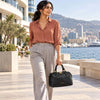 A woman walking along a seaside promenade carrying the black pebbled Italian leather duffle bag with gold-tone hardware. She is wearing a rust-colored button-down shirt and light gray trousers, with a modern coastal city and yachts in the background.