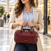 A woman in a beige blazer and light blue shirt holds a bordeaux pebbled Italian leather duffle bag with gold hardware in a bright, modern shopping mall.
