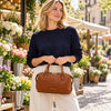 A woman wearing a navy blue sweater and cream trousers stands in a flower market setting, holding the Cognac Italian Leather Duffle Bag with gold-tone hardware.