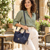 A woman in a green shirt and cream pants carries a dark blue Italian Handbag with gold-tone hardware and a red interior lining, set against an outdoor patio background.