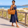 A woman in a navy dress carrying a Italian Leather Tote Bag on a boardwalk near the beach.