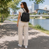 A woman in a casual outfit wearing a the Ladies Backpack while walking by a river in a cityscape.