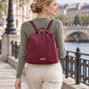 A woman wearing a sage green sweater and cream pants is shown from behind, carrying the Bordeaux Ladies Backpack with gold-tone hardware on a stone bridge in Paris.