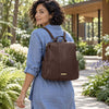 A woman wearing The Chocolate Ladies Backpack with gold-tone hardware, photographed outdoors on a garden path with a modern house in the background.