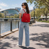 A woman wearing a the Ladies Backpack in red, strolling by a riverside promenade on a sunny day.