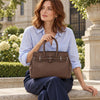 A woman in a light blue shirt and navy pants sits on a stone ledge outdoors, holding The Chocolate Ladies Handbag with top handles and silver-tone hardware.