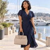 A woman stands on a waterfront promenade wearing a navy wrap-style midi dress with short flutter sleeves and a tied waist, holding Light Taupe Ladies Handbag. Yachts, water, and a sunny outdoor setting are visible in the background.