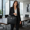 A professional woman in a black suit holding a Ladies Leather Laptop Case in a modern office setting.