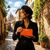 Elegant woman holding an orange Ladies Zipper Wallet in a picturesque street with historical buildings.