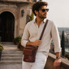 Stylish man wearing sunglasses and beige shirt, carrying Mens Crossbody Bag against a scenic background.