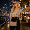 Stylish woman wearing sunglasses and a leather chain clutch bag, posing at a waterfront restaurant at night.