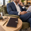 A man in a blue suit sits in an armchair reading a magazine, with The Dark Brown Leather Planner - Weekly Diary on a round wooden table in the foreground. The Leather Planner - Weekly Diary has a light tone tab closure and subtle branding text on the front.