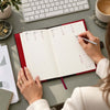 An open lipstick red  Leather Planner - Weekly Diary on a gray desk, with a person's hands writing in it with a silver pen. A keyboard, coffee cup, papers, and small plant are visible around it.
