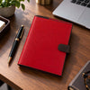A lipstick red Leather Planner - Weekly Diary with a dark brown strap closure, placed on a wooden desk next to a black and gold pen, with a laptop and plant in the background.