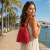 Woman carrying a red leather backpack for women while walking along a waterfront promenade with palm trees.