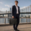 Man in a suit holding a Leather Briefcase Bag by a waterfront with a city skyline in the background.