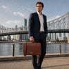 Stylish man holding a Leather Briefcase Bag by the waterfront with a cityscape backdrop.