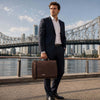 Man in a suit holding a Leather Briefcase Bag with a city skyline in the background near the water.