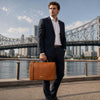 Man in a suit holding a Leather Briefcase Bag near a river with a bridge in the background.