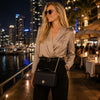 Stylish woman wearing sunglasses and a leather chain clutch bag in black, posing at a waterfront restaurant at night.