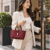 A woman wearing a beige blazer holds the Leather Handbag in a Bordeaux color while walking into a building 