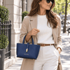 A woman wearing a beige blazer holds the Leather Handbag in a dark blue color while walking down a city street.