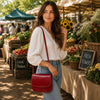 Woman at market holding a Saddle Handbag with red flap, surrounded by fresh produce and flowers.