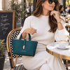 A woman wearing a cream jumper holds the Leather Handbag in a teal color while sitting down at a local cafe