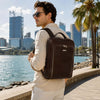 Man wearing a Leather Laptop Backpack near a waterfront with city skyline in the background.