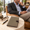 A man in a gray blazer and light blue shirt sits in a leather chair reading, while the Dark Taupe Leather Planner - Weekly Diary rests on a round stone table in the foreground. A black pen and eyeglasses are also on the table, with potted plants and a city view in the background.