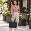 A woman stands on a city sidewalk holding the Black Leather Shopping Tote Bag. She is wearing a dusty pink long-sleeve blouse and light beige trousers, with outdoor cafe seating and flower planters in the background.