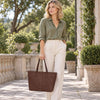 A woman stands outdoors in a sunlit stone walkway wearing an olive green button-up shirt, cream wide-leg trousers, and holding the dark brown Leather Shopping Tote Bag.