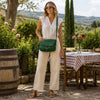 Woman in a white blouse and cream pants holding a green Leather Shoulder Bag in a garden setting.