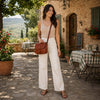 Woman carrying a Leather Shoulder Bag in a charming outdoor setting with a stone wall and table.