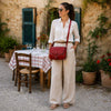 Woman in a stylish white outfit featuring the Leather Shoulder Bag in red, standing in a charming outdoor setting.