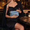 A woman in a black dress holding a stylish blue shoulder bag for ladies at a bar.