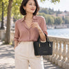 A woman stands outdoors by a waterfront wearing a dusty pink button-up blouse, cream trousers, and carrying The Black Small Leather Tote with gold-tone hardware.