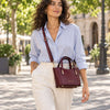 A woman wearing a light blue button-down shirt and cream trousers carries Bordeaux Small Leather Tote as a crossbody bag on a sunny, tree-lined street.