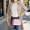 Woman in a beige trench coat carries a Ballet Pink Small Leather Handbag on the street, showcasing elegant style.