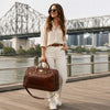 Woman carrying a Small Leather Trolley Bag by the river with a cityscape in the background.