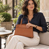 A woman sits on a patio chair holding the Cognac Small Tote Bag.  She wears a dark navy button-up shirt and cream pants, with a coffee cup on a table nearby and plants and stone architecture in the background.