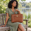 A woman in an olive green dress sits on a wooden park bench holding the Cognac  structured leather tote bag , set against a blurred background of a lake and green trees.