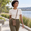 A woman walking outdoors on a lakeside path wearing a cream short-sleeve knit top, olive green pants, and The Dark Taupe Wallet And Phone Holder