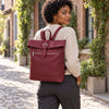 A woman wearing a cream ribbed sweater and dark gray trousers carries The Bordeaux Womens Leather Backpack on her back while walking down a cobblestone street lined with brick buildings and green ivy.