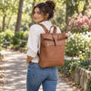 A woman wearing a white blouse and blue jeans is shown from behind on a park path, carrying The Cognac Womens Leather Backpack. The backpack has a flap top with a buckle strap, a front zip pocket, and silver hardware.