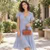 A woman wearing a light blue ruffled midi dress with short flutter sleeves and a V-neckline, holding The Cognac Womens Leather Clutch in an outdoor garden courtyard setting.